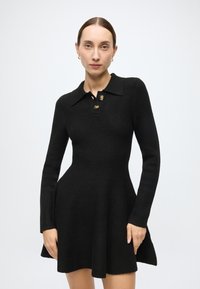 Black dress with long sleeves, a collar, and three gold buttons at the neck. Fitted torso flares into a flared skirt. Textured fabric.