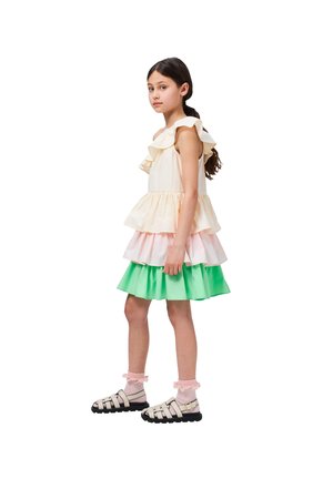 Πολύχρωμο φόρεμα με επίπεδα και ρυάκια στις τιάρες; ανοιχτό κίτρινο επάνω μέρος, παστέλ ροζ και πράσινα στρώματα. Μαύρες σανδάλες με λουριά και ροζ κάλτσες.