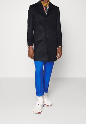 Personne portant un manteau noir boutonné, un pantalon bleu retroussé aux chevilles, des baskets blanches et un pull coloré à motifs visible sous le manteau.