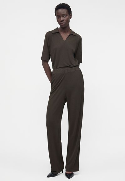 Γυναίκα όρθια με τα χέρια στις τσέπες, φοράει σκουρόχρωμο ελιά κοντομάνικο πουκάμισο με γιακά και ταιριαστό παντελόνι με φαρδιά μπατζάκια, μαζί με μαύρα μυτερά παπούτσια.