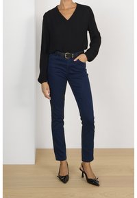 Femme portant un blouse noire à col en V à manches longues, un jean slim bleu foncé avec une ceinture noire, et des chaussures à talons noirs à bout pointu et à bride arrière.
