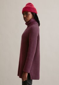 Gebreide coltrui in diep paars met een gestructureerd patroon, gecombineerd met een heldere roze, zachte beanie, zijaanzicht, met zichtbare zijsplitten.