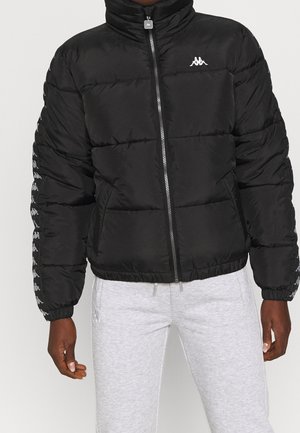 Personne portant une doudoune noire avec un logo blanc sur la poitrine et le long des manches, associée à un pantalon de survêtement gris clair.