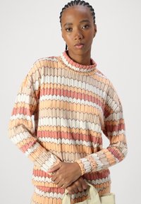 Flerfarget genser med ribbet tekstur og zigzag-mønster i fersken, oransje, krem og rustnyanser. Mock turtleneck og lange ermer.