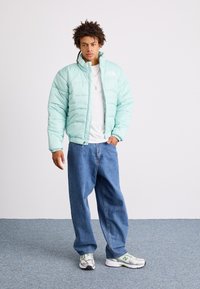 Giacca puffer verde menta con chiusura a zip, polsini a coste, indossata sopra una maglietta bianca. Abbinata a jeans blu larghi e sneakers grigie.
