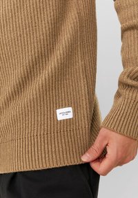 Brun ribbad stickad tröja med en etikett vid fållen. Har en texturerad design och åtsittande manschetter, tillverkad av mjukt material.