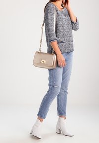 Grau strukturiertes Sweatshirt mit Dreiviertelärmeln, kombiniert mit hellblauen Ripped Jeans und weißen Knöchelstiefeln. Hält eine metallisch goldene Handtasche.