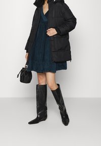 Musta toppatakki hupulla, turkoosi puuvillafroteinen mekko, mustat nahkaiset polven yli ulottuvat saappaat ja pieni musta laukku.