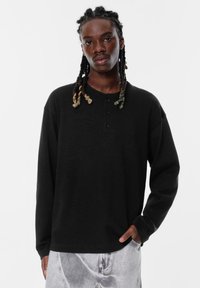 Schwarzes Langarm-Henley-Shirt mit weicher Textur, ausgestattet mit einer Knopfleiste mit drei Knöpfen und lockerer Passform, kombiniert mit hellgrauen Jeans.