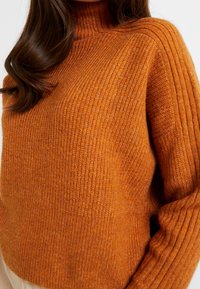 Pomarańczowy sweter na drutach z wysokim kołnierzem, z prążkowaną fakturą na ramionach i rękawach, podkreślający ciepło i przytulny design.