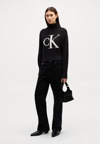 Maglione nero a coste con collo alto caratterizzato da un prominente logo "CK" bianco, abbinato a pantaloni neri a gamba larga e una piccola borsa nera.