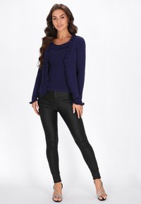 Marineblauer gerippter Pullover mit Rüschensaum, kombiniert mit schwarzen leggings aus lederähnlichem Material und silbernen Riemchenheels. Modell steht vor einem weißen Hintergrund.