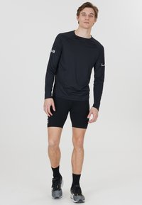 Zwart sportshirt met lange mouwen en ronde halslijn, met raglanmouwen. Gedragen met zwarte shorts en grijze sportschoenen.