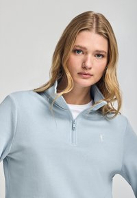 Light blue zip-up sweatshirt made of soft fabric, featuring a high collar and small embroidered logo on the chest. Smooth texture.