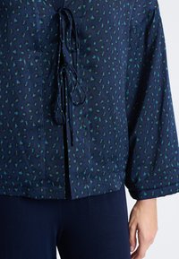 Granatowa bluzka w zielone panterki; posiada zapięcie z przodu na sznur, długie rękawy oraz luźny krój. Miękka tkanina o gładkiej fakturze.