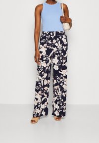 Jasnoniebieski top bez rękawów w połączeniu z granatowymi spodniami w kwiaty o szerokich nogawkach, zdobionymi białymi i różowymi kwiatami, dopełniony beżowymi sandałkami i małą torebką.