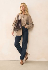 Smėlio spalvos lininiai marškiniai, tamsiai mėlynos spalvos aukštakulniai džinsai su aukštu liemeniu, rudi zomšiniai smailūs aukštakulniai ir ruda odinė rankinė, ant neutralaus fono.