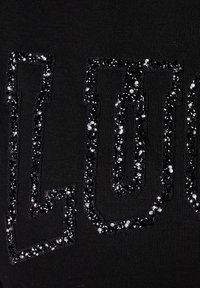 Un tissu noir présente de grandes lettres dessinées, ornées de brillants strass noirs et argentés, créant un contraste texturé avec le matériau.