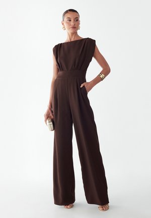 Kvinde iført ærmeløs, vidbenet brun jumpsuit, holder lille metallisk clutch, accessoriseret med gyldne øreringe og armbånd, kigger mod venstre.