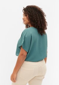 Zielona bluzka z krótkimi rękawami o luźnym kroju, teksturowana tkanina, okrągły dekolt i proste wykończenie, w połączeniu z jasnobeżowymi dopasowanymi spodniami.