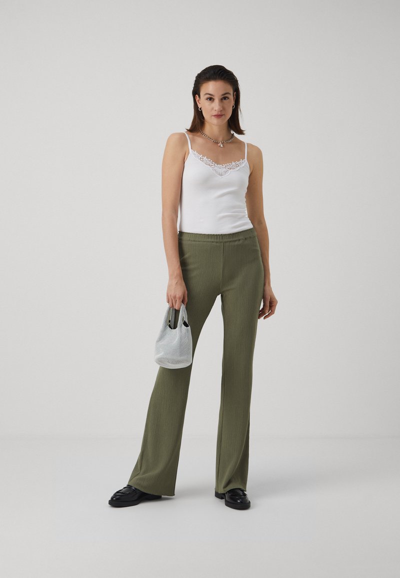 Débardeur blanc avec dentelle, pantalon large côtelé vert olive, mocassins noirs et un petit sac à carreaux gris. Tenue simple et décontractée.