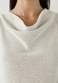 Close-up of a person wearing a white knitted cowl neck sweater, showing the neckline and textured fabric.