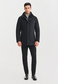 Czarny ocieplany płaszcz z wysokim kołnierzem, z zatrzaskami i gładką fakturą. W zestawie z czarnymi spodniami i eleganckimi butami.