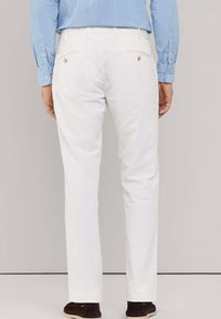 Hombre que lleva pantalones blancos y zapatos marrones, mostrado desde atrás contra un fondo neutro liso.