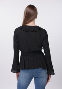 Blouse noire avec encolure à volants, manches longues évasées et taille smockée. Associée à un jean bleu clair ajusté. Texture de tissu lisse.