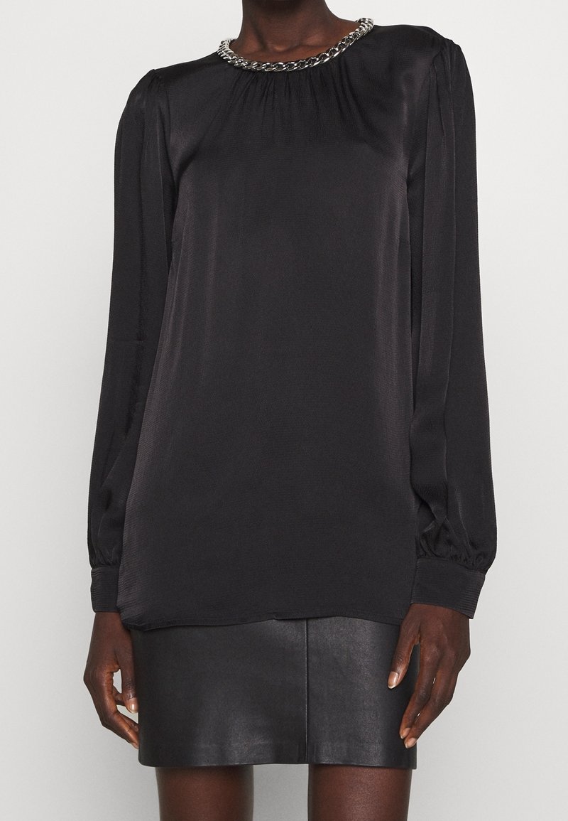 Blouse noire à manches longues avec détail de col en chaîne argentée, associée à une mini-jupe en cuir noire portée par un modèle debout.