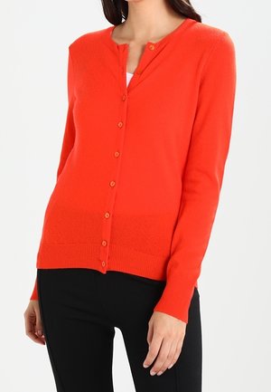 Femme portant un cardigan orange vif boutonné à manches longues et un pantalon noir, se tenant devant un fond clair uni.