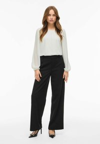 Blusa bianca a maniche lunghe abbinata a pantaloni neri a vita alta e gamba larga, con tacchi a punta neri decorati con dettagli in catena dorata.