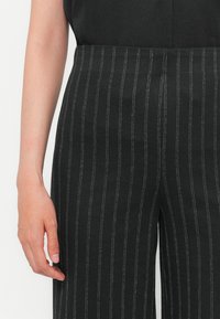 Pantalons rayés noirs avec un tissu texturé et des rayures blanches, coupe fuselée et taille haute. Main positionnée près de la taille.