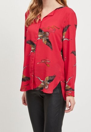 Blusa roja de manga larga con estampados de grullas en negro, blanco y dorado, combinada con pantalones negros, mostrada desde la mitad del torso hasta las caderas.