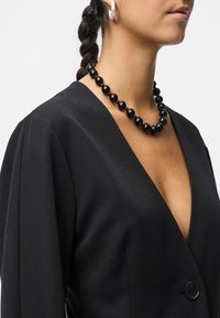 Černý top s výstřihem do V a strukturovaným siluetou, s jedním knoflíkem na zavírání a záhyby na ramenech. Doplněn náhrdelníkem z černých korálků.