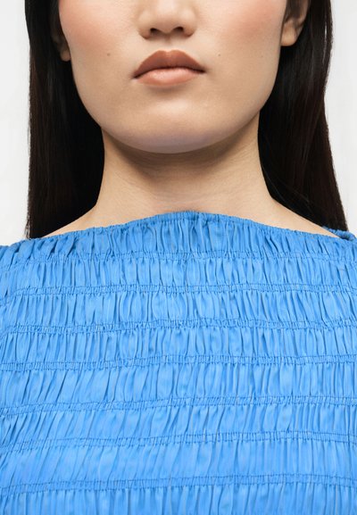 Κοντινό πλάνο του κάτω μέρους του προσώπου και του λαιμού ενός ατόμου που φοράει υφασμένο, ανοιχτό μπλε μπλουζάκι με ακάλυπτους ώμους και έχει μακριά σκούρα μαλλιά.