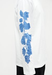 Λευκό πουκάμισο με μακριά μανίκια και μπλε μοτίβο με βοτανικό κλήμα κατά μήκος του εξωτερικού μανικιού, φορεμένο από ένα άτομο που στέκεται μπροστά σε απλό φόντο.