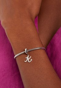 Bracelet en argent avec un design texturé, comportant un charm en forme de "K" suspendu. Porté sur un poignet contre un fond texturé rose.