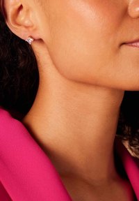 Boucle d'oreille en or avec une pierre précieuse carrée et transparente. Gros plan sur un teint de peau et un tissu rose. Texture lisse avec un éclat naturel.
