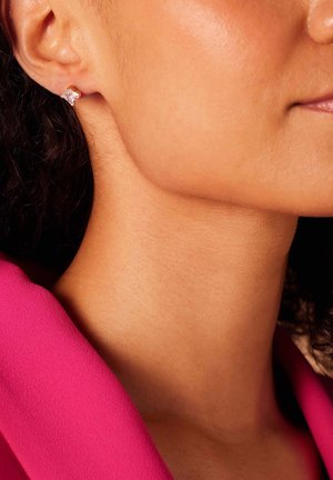 Boucle d'oreille en or avec une pierre précieuse carrée et transparente. Gros plan sur un teint de peau et un tissu rose. Texture lisse avec un éclat naturel.