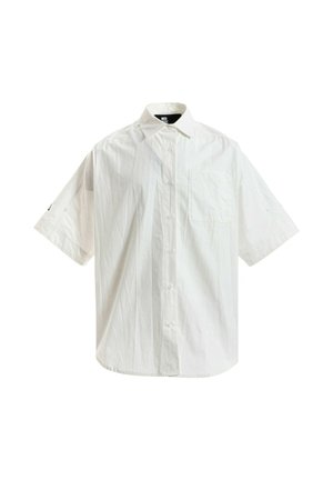 Joven con rastas cortas lleva una camisa blanca de manga corta y muy grande, pantalones anchos beige con cadena y zapatos marrones de plataforma, está quieto.