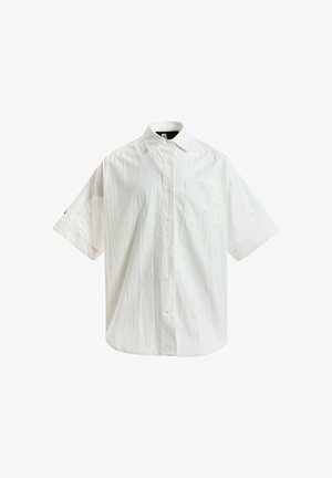 Joven con rastas cortas lleva una camisa blanca de manga corta y muy grande, pantalones anchos beige con cadena y zapatos marrones de plataforma, está quieto.