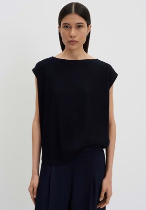 Femme aux cheveux lisses et foncés porte un haut noir texturé sans manches et un pantalon sombre, se tenant devant un fond clair uni.