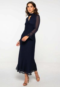 Vestido de manga larga azul marino con escote en V, cuerpo arrugado y mangas de chifón acampanadas. Acentos de tela transparente en los puños y el dobladillo.