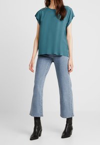 Blouse turquoise à manches courtes avec une coupe décontractée, associée à un jean évasé bleu clair et des bottes à talons noires, présentant une texture lisse et un design simple.