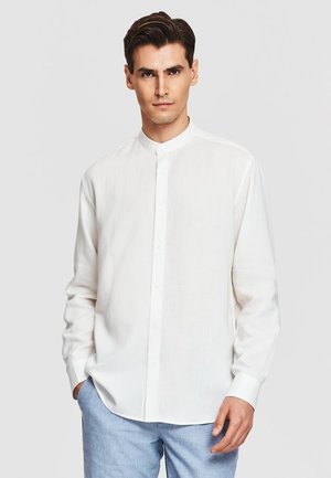 Mężczyzna w białej koszuli z długim rękawem i kołnierzykiem mandaryńskim oraz jasnoniebieskich spodniach, stojący z jedną ręką w kieszeni na jednolitym tle.