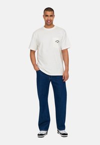 Witte katoenen t-shirt met een borstzak en zwarte tekst, gecombineerd met donkerblauwe spijkerbroek en zwart-witte sneakers.