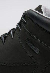 Botín negro de cuero con lados texturizados, costuras en contraste y un parche con el logo. Presenta cordones redondos y ojales metálicos para asegurar.