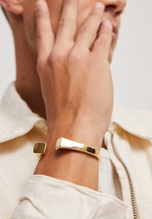 Bracelet en or avec une surface lisse et brillante, présentant des extrémités géométriques : une forme carrée et une forme rectangulaire, sur un fond neutre.