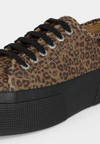 Αθλητικό παπούτσι με καφέ και μαύρο ύφασμα με λεοπάρ σχέδιο, μαύρη παχιά υφή στην πλατφόρμα της σόλας και μαύρα κορδόνια που περνούν μέσα από ασημένια λογχοειδή.